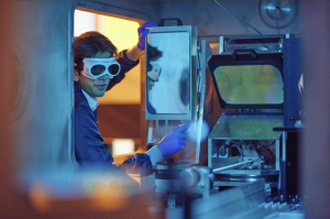 scientist with laser goggles peers into the laser target chamber. The chamber is chrome and mirrored, and the scene is bathed blue and orange light.