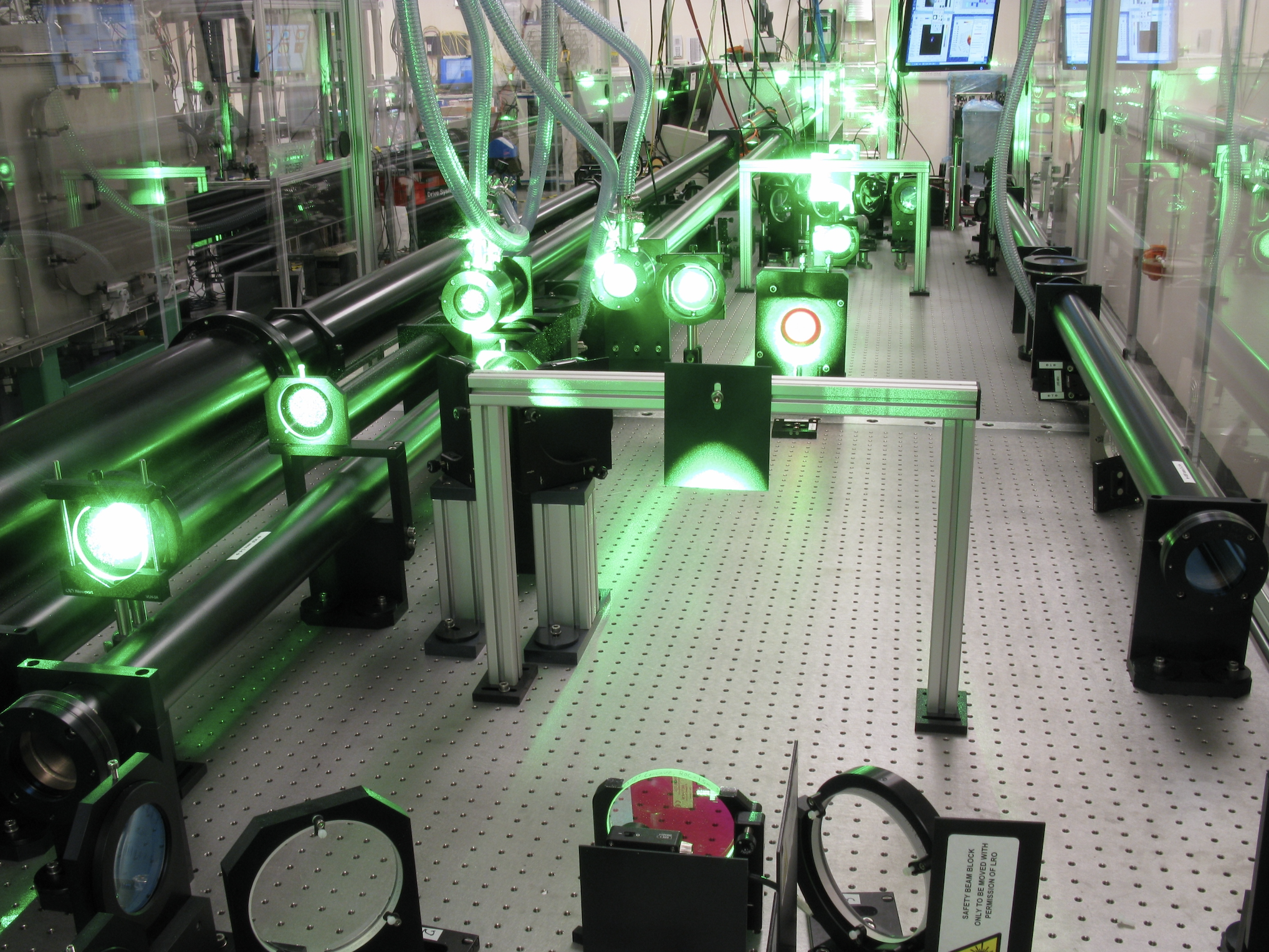 A metal table with several different types of mirrors and lenses bolted to it. There is a bright green laser passing through it, which can be seen as it shines on each mirror or lens.