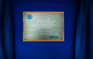 Metal commemorative plaque mounted in a wooden frame reading: “This building for the Extreme Photonics Applications Centre is dedicated to the memory of Professor David Neely, an inspirational CLF scientist without whom it would not have been possible,” with Central Laser Facility and UKRI Science and Technology Facilities Council logos.